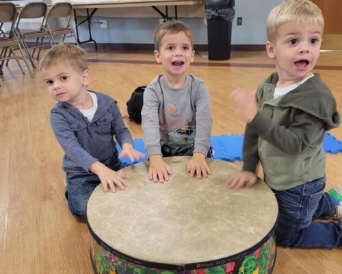 Three young children gather around a large drum during a music class.