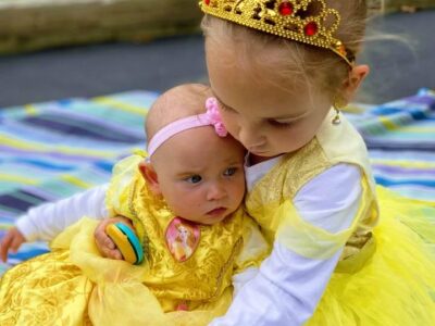 Older child holding baby during outdoor music and movement class. Both children are playing castanets and are dressed as Belle.