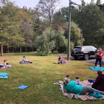Outdoor Music Class for Kids and Parents Ages birth through 4. Teacher playing guitar while families sit on blankets in a circle singing along.